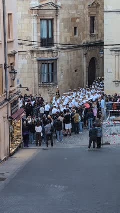 Crowd gathers on a narrow street in front of historic stone buildings, some participants in white robes heading toward an archway.